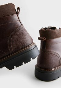 Botas de cuero marrón hasta el tobillo con paneles texturizados, puntera redonda y suela de goma con un diseño de perfil. Incluyen una cinta en la parte trasera.