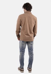 Camicia marrone con colletto, maniche lunghe e due spalle posteriori, abbinata a jeans blu sbiaditi e sneaker con motivo.