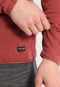 Maglietta a maniche lunghe di colore marrone rossastro, realizzata in tessuto strutturato, con una piccola etichetta nera con la scritta "ONLY & SONS" vicino all'orlo.