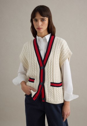 Vrouw draagt een crème kabelgebreid vest met rode en marineblauwe bies over een wit overhemd en donkere broek, staand tegen een effen achtergrond.