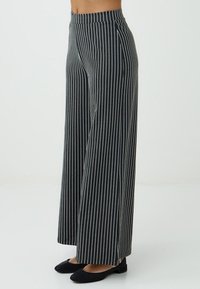 Vrouw in hoogstaande, wijde zwarte broek met witte strepen, gecombineerd met zwarte flats, staand in profiel tegen een effen achtergrond.
