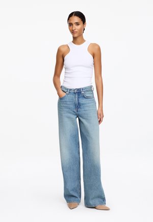 Kobieta z zaplecionymi włosami, ubrana w białą, bezrękawnikową bluzkę, jasnoniebieskie, szerokie spodnie o wysokim stanie oraz beżowe, spiczaste buty, stoi z jedną ręką w kieszeni.