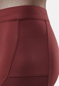 Hoge sportleggings in bordeauxrood. Soepele stof met zichtbare stiksels, zijzak en een textuurpaneel op het onderbeen.