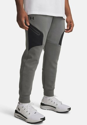 Man die grijze joggingbroek met zwarte panelen en witte sportschoenen draagt, staat met ontspannen armen langs zijn zij.