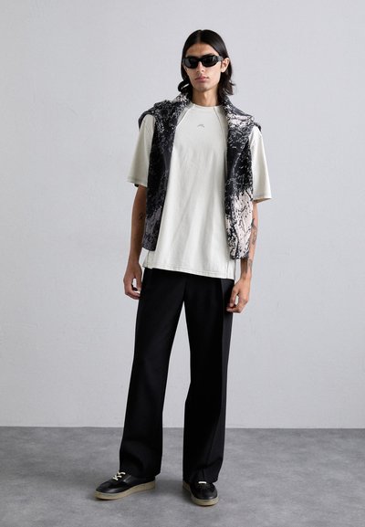 Férfi öltözet, amely egy könnyű, oversized tojássárgája színű pólóból és egy ujjatlan, fekete és szürke mintás mellényből áll. Fekete, bő szárú nadrág és napszemüveg kíséri.