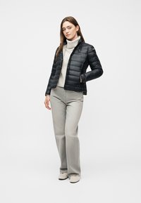 Femme portant une doudoune noire par-dessus un col roulé blanc, un pantalon gris à jambes larges et des chaussures de couleur claire, debout avec une main dans la poche.