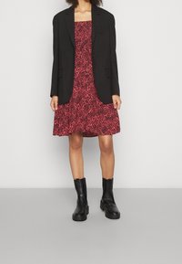Schwarze Blazer über einem roten Kleid mit Rosenmuster, kombiniert mit schwarzen Knöchelstiefeln. Das Kleid hat ein tailliertes Oberteil und einen ausgestellten Rock.