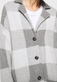 Manteau en laine à carreaux gris et blanc avec des boutons noirs, porté par-dessus un t-shirt blanc à col rond.