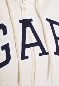 Sudadera cremosa con cremallera que presenta un gran bordado en azul marino de "GAP" en el frente con contorno blanco. Incluye detalles de cordón a juego.