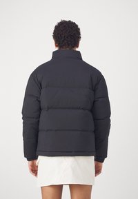 Osoba z kręconymi włosami, ubrana w czarną puchową kurtkę i białą spódnicę, pokazana od tyłu na jednolitym tle.