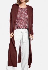 Cardigan lungo borgogna, aperto davanti, realizzato in tessuto morbido. Indossato sopra una maglietta rossa e bianca a fantasie e pantaloni bianchi a gamba larga con cintura.