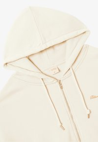 Cremefarbener Kapuzen-Zip-Hoodie mit flauschigem Innenfutter. Verfügt über einen silbernen Reißverschluss und ein gesticktes Logo auf der Brust. Weicher, gemütlicher Stoff.