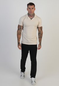 Beige poloshirt met kraag, korte mouwen; gecombineerd met zwarte jeans en witte sneakers, die een getailleerde stijl en tattoos tonen.