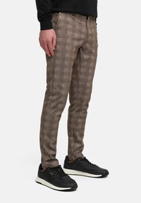 Figura masculina con pantalones marrones estampados y zapatillas negras, de pie con la mano restingando en la pierna, contra un fondo blanco liso.