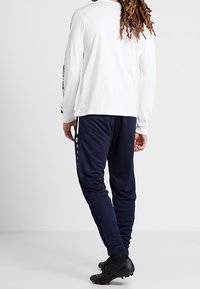 Osoba s dlouhými dredy má na sobě bílou košili s dlouhým rukávem a tmavě modré sportovní kalhoty s knoflíky na boku, a černé kopačky, stojí na šedé podlaze.