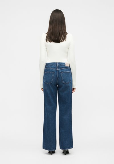 Γυναίκα με ίσια καστανά μαλλιά, που φοράει λευκό μακρυμάνικο τοπ και μπλε τζιν με ψηλή μέση και φαρδιά πόδια, με την πλάτη προς τα εμπρός σε λευκό φόντο.
