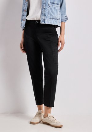 Person trägt schwarze, kurze Cargohosen, beige Sneakers, weißes Hemd und hellblaue Jeansjacke, steht vor einem schlichten hellen Hintergrund.