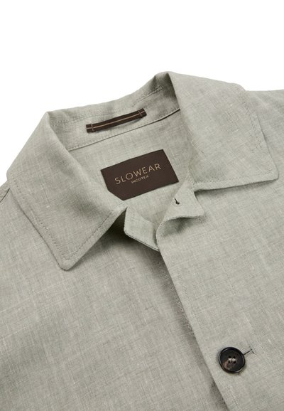 Jasnoszara lniana koszula z ostrokątnym kołnierzem, ciemnym guzikiem u dołu oraz metką z napisem "SLOWEAR INCOTEX" wewnątrz kołnierza.