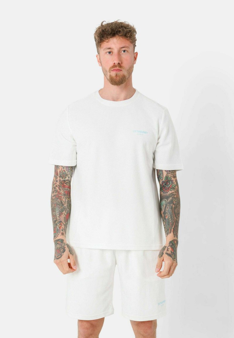 Weißes Baumwoll-T-Shirt mit kurzen Ärmeln und Rundhalsausschnitt, mit einem hellblauen Logo auf der Brust. Kombiniert mit passenden weißen Shorts.