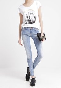 Camiseta blanca de manga corta con un gráfico del horizonte de Londres, combinada con jeans ajustados de tiro bajo en azul claro y zapatos negros con cordones. Sosteniendo un clutch negro.