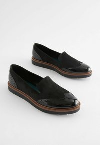 Scarpe slip-on nere in una combinazione di camoscio e pelle verniciata, con dettagli brogue, suola piatta e un accento marrone a contrasto.