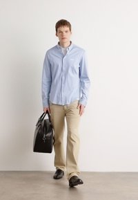 Lichtblauw blouse met lange mouwen en geborduurd logo, beige broek, zwarte schoenen en een zwarte leren tas met een zilveren accent.