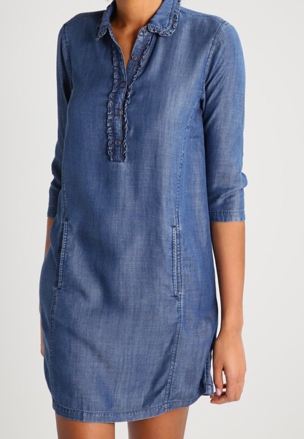 Jeansblå klänning med 3/4-långa ärmar, volangkrage, knäppt plagg och sidofickor, bärs av en person med ena handen avslappnat vid sidan.