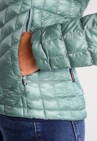 Prošívaná světle tyrkysová bunda z lesklé látky, vybavená kapsou na zip a elastickými manžetami pro těsné přilnutí. Viditelné vzory stehu.