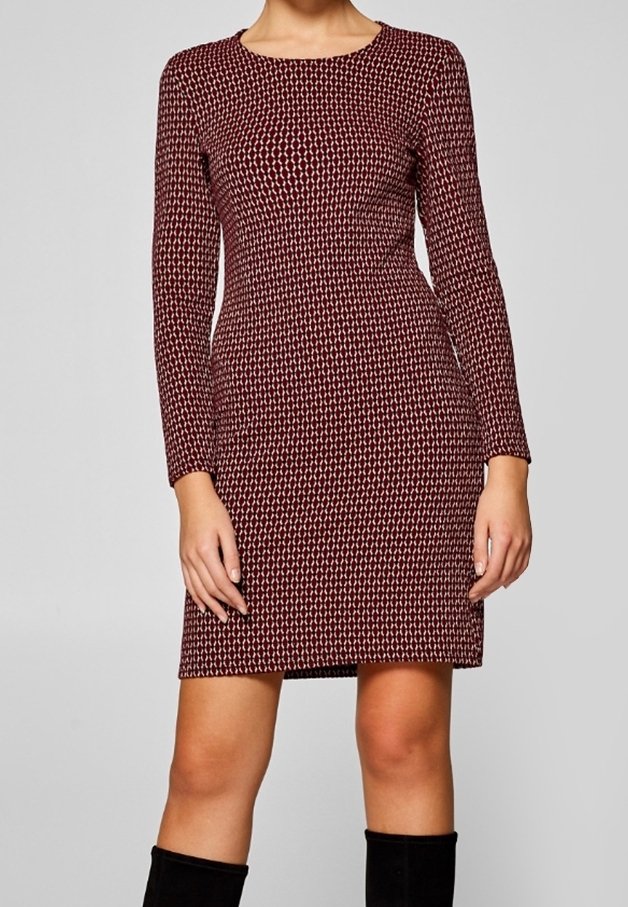 Femme portant une robe imprimée rouge et blanche, longue à manches longues jusqu'aux genoux, avec des bottes noires montantes, debout devant un fond uni.