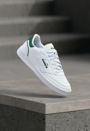 Valge nahast tennisking rohelistes detailides. Omab perforeeritud varbaosa, nööritavat eesosa ja tekstuurse kummist talla. Olevasti hõljuv betoonpinnal.