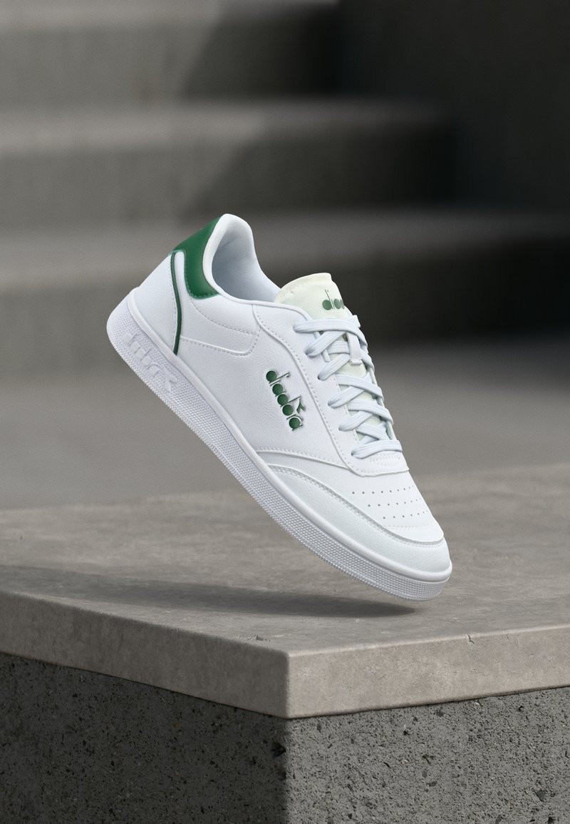 Sneaker in pelle bianca con dettagli verdi. Presenta punta traforata, chiusura con lacci e suola in gomma texturizzata. Fluttua sopra una superficie di cemento.