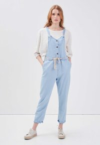 Salopette in denim azzurro chiaro con spalline regolabili, chiusura a bottoni, tasche laterali e una cintura intrecciata. Abbinata a un maglione larghi in maglia color crema.