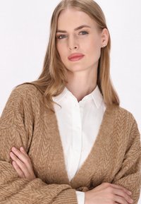 Cardigan bej tricotat, cu decolteu în V, model texturat cu cabluri și mâneci largi, purtat peste o cămașă albă cu guler.