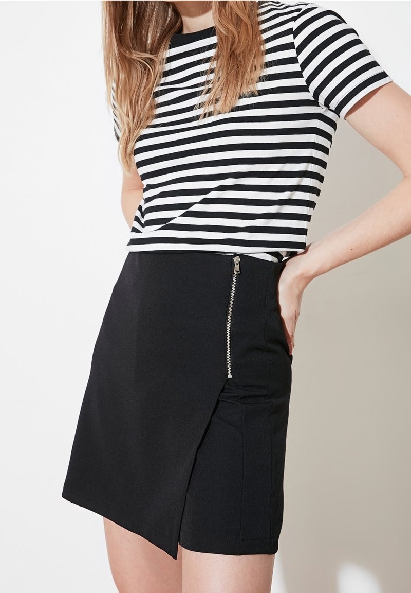 Femme portant une jupe asymétrique noire avec une fermeture éclair latérale et un t-shirt rayé horizontal noir et blanc, main posée sur la hanche.