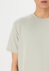 Ανοιχτοπράσινη υφασμάτινη μπλούζα T-shirt με τετράγωνο σχέδιο, στρογγυλή λαιμόκοψη και κοντά μανίκια. Το ύφασμα φαίνεται μαλακό και ελαφρώς ελαστικό.