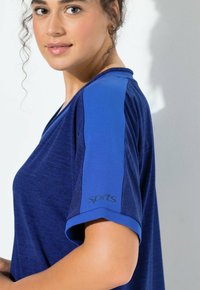 Młoda kobieta ubrana w niebieską sportową koszulkę z jaśniejszym niebieskim paskiem na rękawie, zwrócona częściowo bokiem na białym tle.