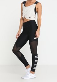 Femme portant un legging Nike noir avec des panneaux en mesh et le texte « JUST DO IT. », des chaussures de sport blanches et un haut blanc sans manches noué à l'avant.