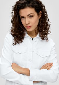 Witte denimjas met een knoopsluiting aan de voorkant, twee borstzakken en lange mouwen. Krullend haar gedeeltelijk zichtbaar. Achtergrond is neutraal.