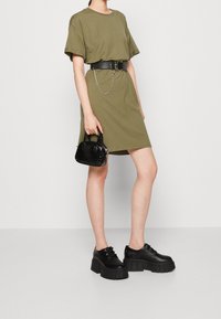 Sukienka t-shirt w oliwkowym kolorze o luźnym kroju, czarny skórzany pasek z detalami łańcuszka, czarna torebka z lakierowanej skóry, masywne czarne buty na platformie.