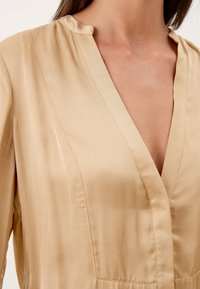 Blouse en satin beige avec un col en V et un col décontracté. Texture lisse avec un léger éclat et des coutures вертикals. Pas de décorations visibles.