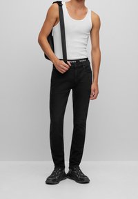 Vaqueros negros con corte ajustado, una cinturilla de Hugo, combinados con una camiseta sin mangas blanca y zapatillas negras, llevando un bolso de hombro negro.