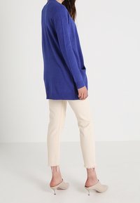 Lång blå stickad kofta med ribbat avslut, bärs över ljusbeiga korta byxor. Mjuk textur, avslappnad passform och sidofickor synliga.