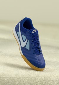 Modra športna obutev Nike z belim logotipom swoosh, modrimi vezalkami in gumijastim podplatom, prikazana na mehkem bež teksturiranem ozadju.