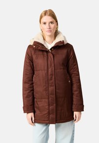 Ruskean eristetehty parka-takki, jossa on tekoturkista vuorattu huppu, vetoketju ja napitus, sekä sivutaskut; takissa on sileä ulkopinta.