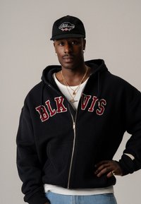 Schwarze Zip-Up-Hoodie mit "BLK VIS" in weinroter Schrift, getragen über einem weißen Hemd, kombiniert mit einer schwarzen Mütze mit gesticktem Design.
