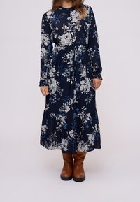 Robe longue à manches longues bleu marine à fleurs, avec une taille cintrée, un ourlet à volants et des motifs de fleurs bleues et blanches. Portée avec des bottes marron montant jusqu'aux genoux.