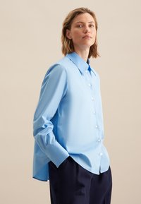 Camicia button-up azzurro chiaro con colletto, maniche lunghe e orlo posteriore leggermente più lungo. Tessuto liscio con una sottile texture.