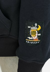 Černá mikina s texturovaným žebrovaným lemem a tkanou nášivkou znázorňující šálek kávy s detaily "CENTRAL PERK" a "F.R.I.E.N.D.S.".