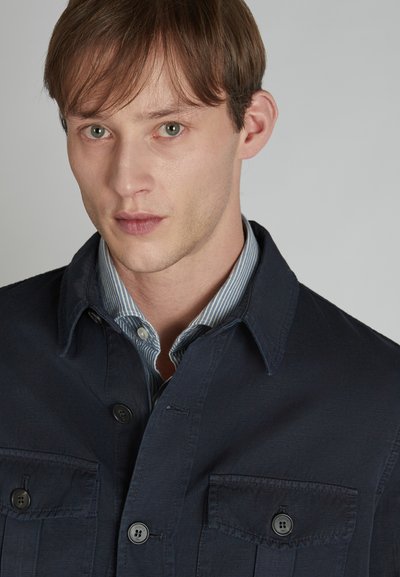 Chemise bleu marine à boutons avec deux poches poitrine, dotée d'un col et d'un tissu texturé. En dessous, une chemise rayée bleu clair est visible.
