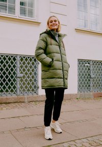 Kārdināta olīvu zaļa gara mētelis ar rāvējslēdzēju un kapuci, ar tekstūrētu apdari un sānu kabatām. Nēsāts ar melnām biksēm un baltām kedām.